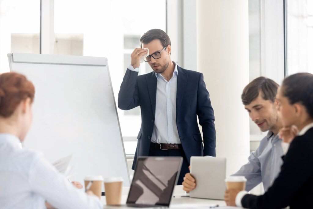 Das Coaching zur Potenzialentfaltung hilft Ihnen Situationen wie auf dem Bild zu vermeiden, wo ein Mann in einem Meeting vor anderen Mitarbeitern reden muss und sich den Angstschweiß von der Stirn wischt.
